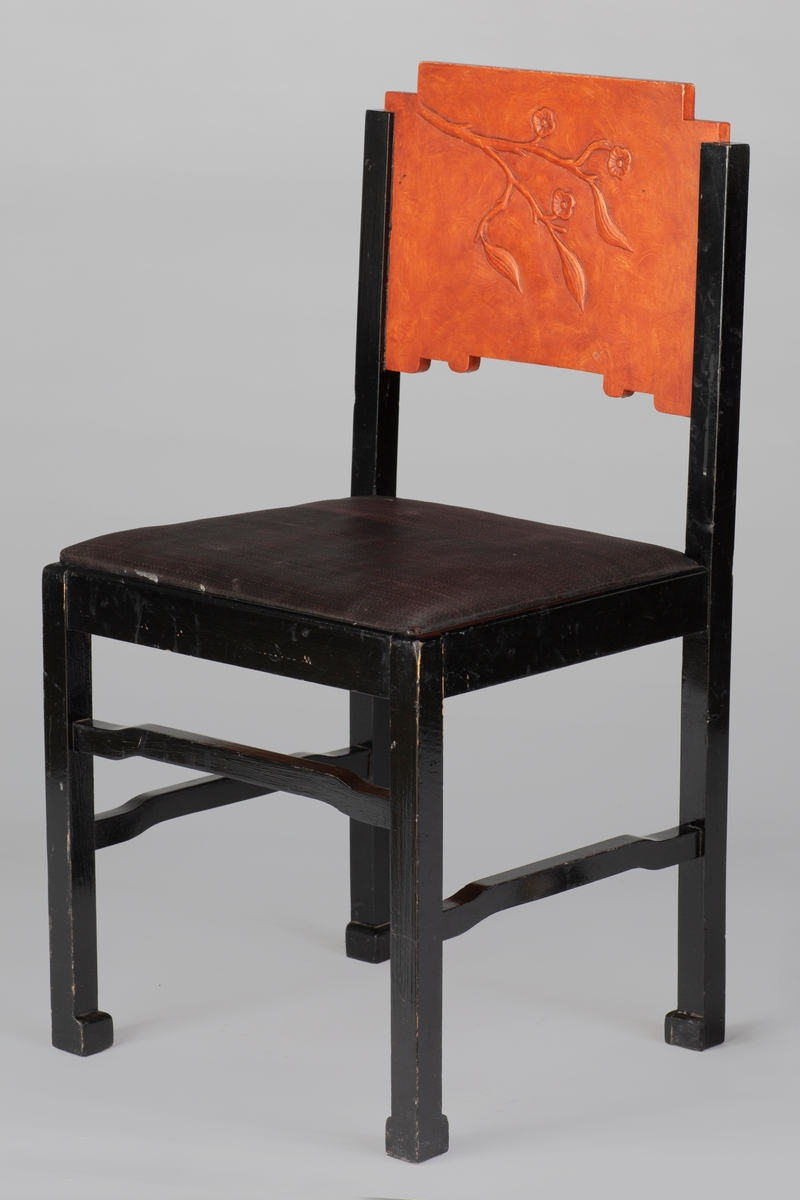 Sortmalt stol i furu med oransj ryggbrett. Ryggbrettet har lærimiterende bemaling og et blad/blomstermotiv skåret i relieff. Løst trapesformet sete med hestehårstrekk.