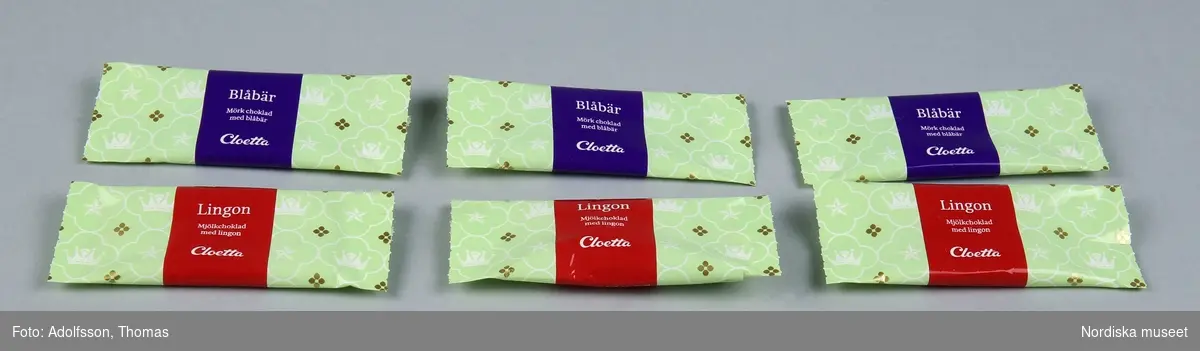 a-f) 6 st plastomslag för chokladbitar. Grön bottenfärg med samma mönster som asken (kronor och Nordstjärnan inskrivna i fyrpass på grön botten).  På tre av dem lila tvärband med text "Blåbär / Mörk choklad med blåbär / Cloetta / 10 g" samt symboler för att plasten kan återvinnas. På tre av dem rött tvärband "Lingon / Mjölkchoklad med Lingon / Cloetta / 10 g" samt symboler för att plasten kan återvinnas. 
/Leif Wallin 2015-01-12
