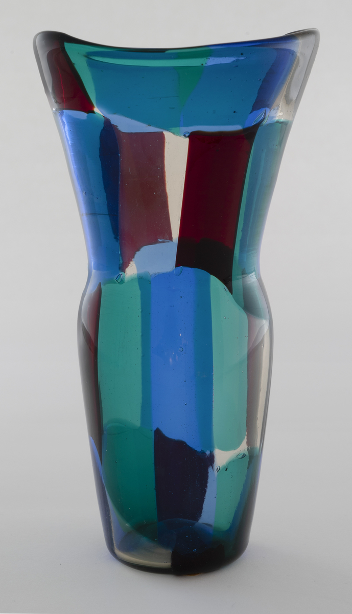 Vase i gjennomskinnelig glass. Korpus er dekorert med rektangulære felter i grønt, rødt, blått og klart glass. Ujevn ovalformet munning og lang konisk hals.