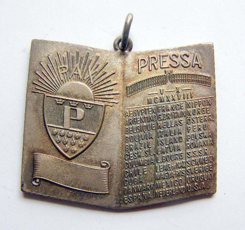 Minnesplakett av silver med ring för upphängning, från den internationella pressutställningen Pressa 1928. Plaketten är formad som ett bokuppslag, med ett vapen och texten PAX på vänstra sidan, och namnet PRESSA samt en lista över förmodade utställare på högra sidan, Sverige bland dem.