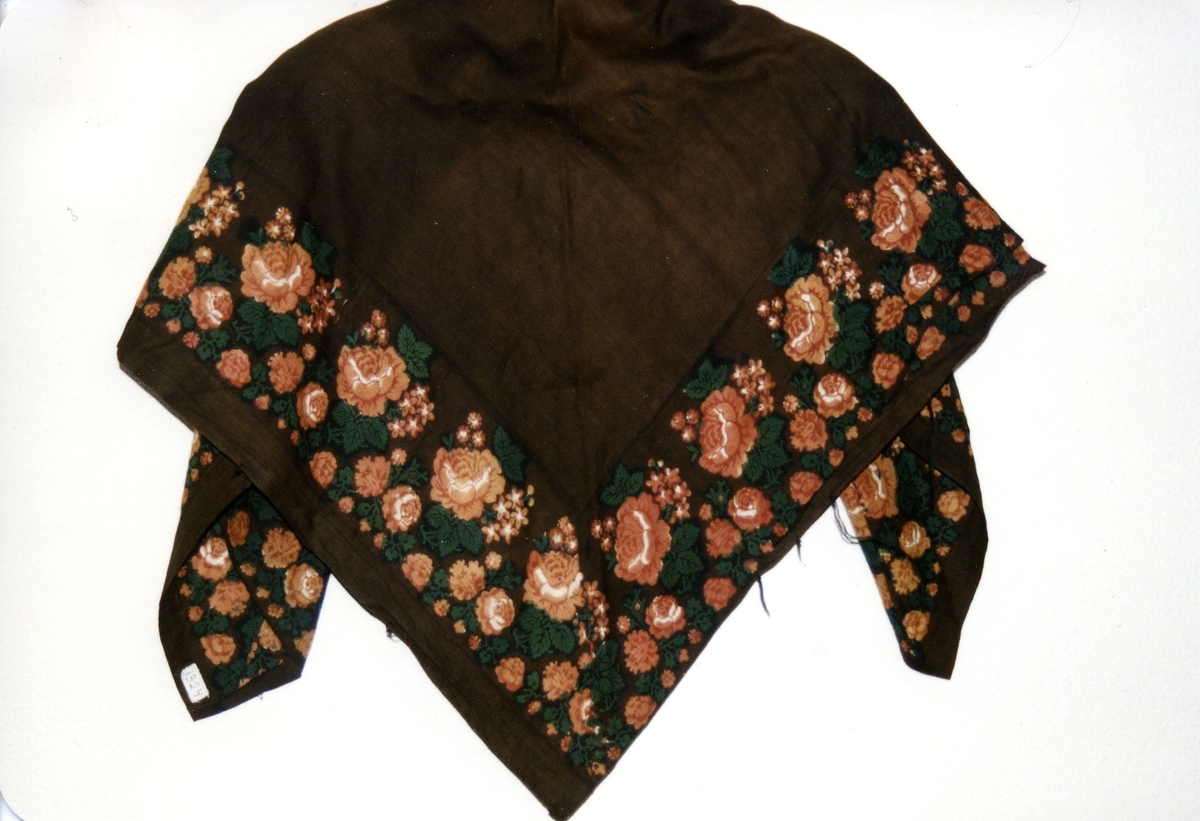 Brunt/svart sjal. Laget av et brun/svart bomullstøy med trykte roser i grønt, kvitt, rose/lys-brun. Merker etter utdradde frynser i ull. 