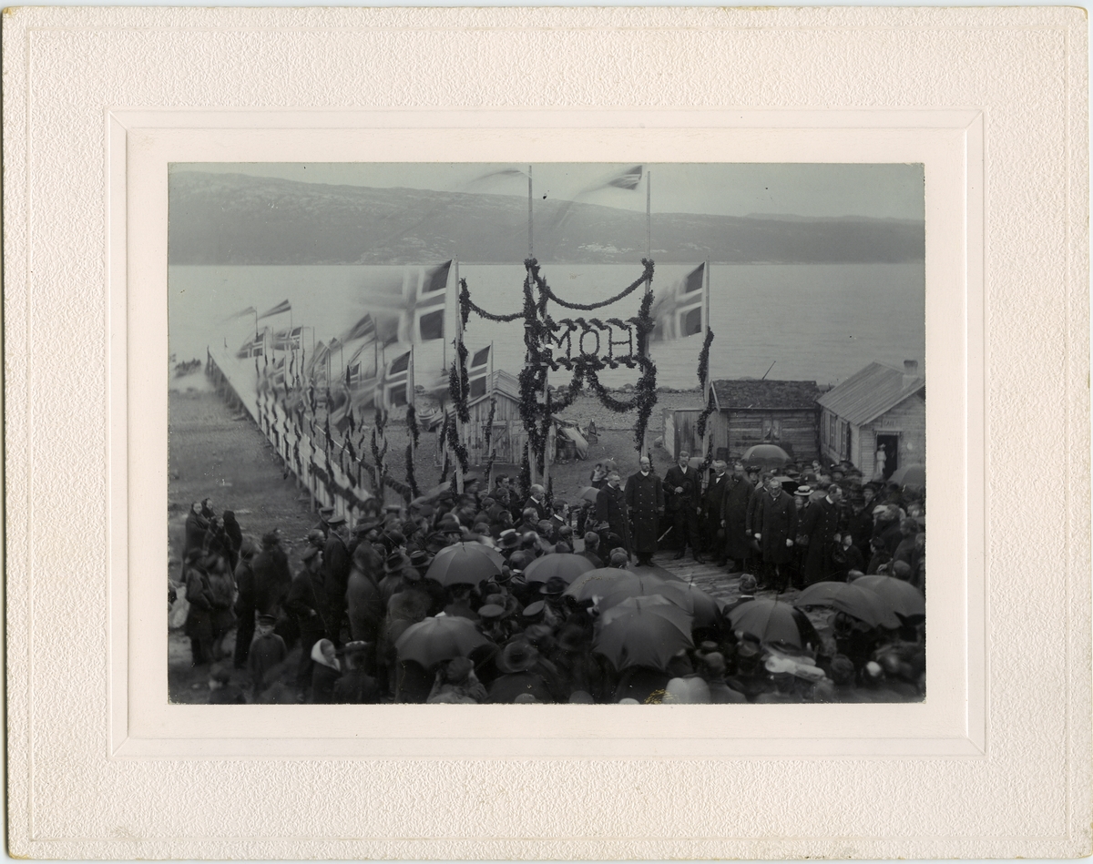Kong Haakon VII og dronning Mauds besøk i Kirkenes 27.07.1907. Vi ser kaia pyntet med flagg og befolkningen er festkledt i anledningen. Kong Haakon hilser på folket.