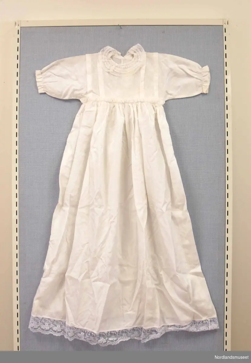 Dåpskjole i hvitt glatt stoff med blomstermønster. Blonder rundt hals og nede på kjolen. Det følger også med rosa og blå silkebånd.