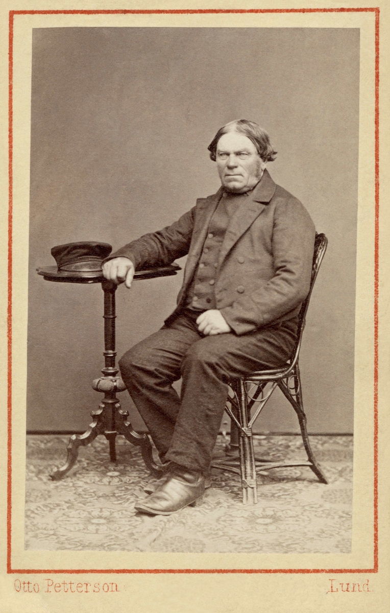 En man sitter på en stol, lutar armen mot ett bord, där han lagt sin vegamössa