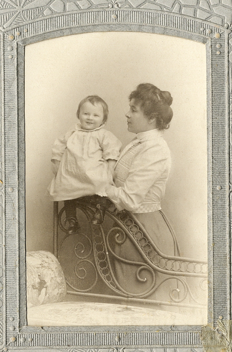 En kvinna i ljus blus och kjol, som håller en glad baby, iklädd kolt.
Knäbild. Ateljéfoto.

Fotografen med dotter.