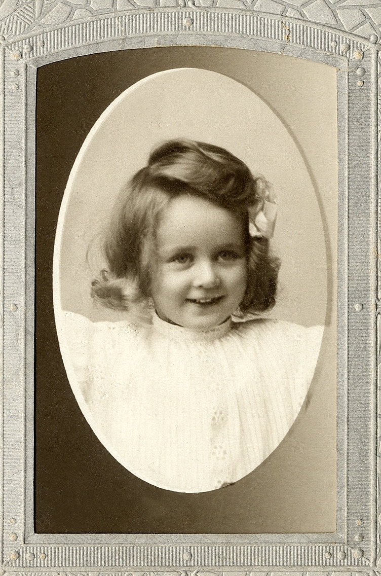 En liten flicka i ljus klänning, med rosett i håret. 
Bröstbild, en face. Ateljéfoto.

Fotografens dotter.
