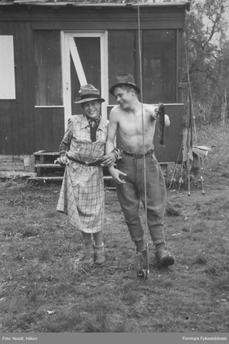 Erik og Ivar Noodt har det moro med dameklær og fiskestang. Ivar holder en fisk, kanskje ørret, i hånda. Bildet er tatt utenfor Leif Noodts hytte i Stabbursdalen i Porsanger, 1954.