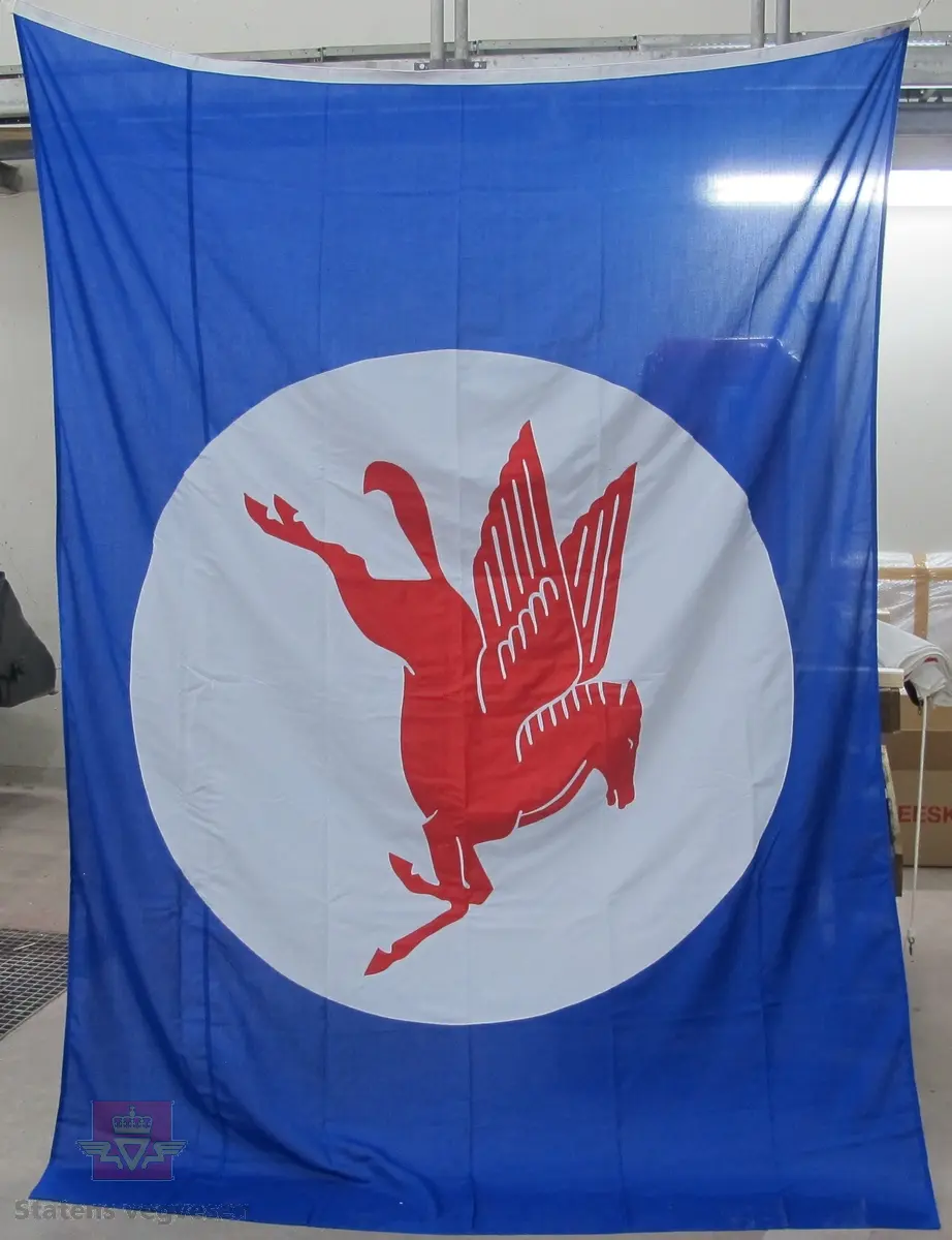 Rektangulært flagg i tekstil med tau og to opphengskroker i plast. Blått med hvit kant på den ene siden og hvit sirkel med Pegasus. Opphengstauet og krokene er hvite.