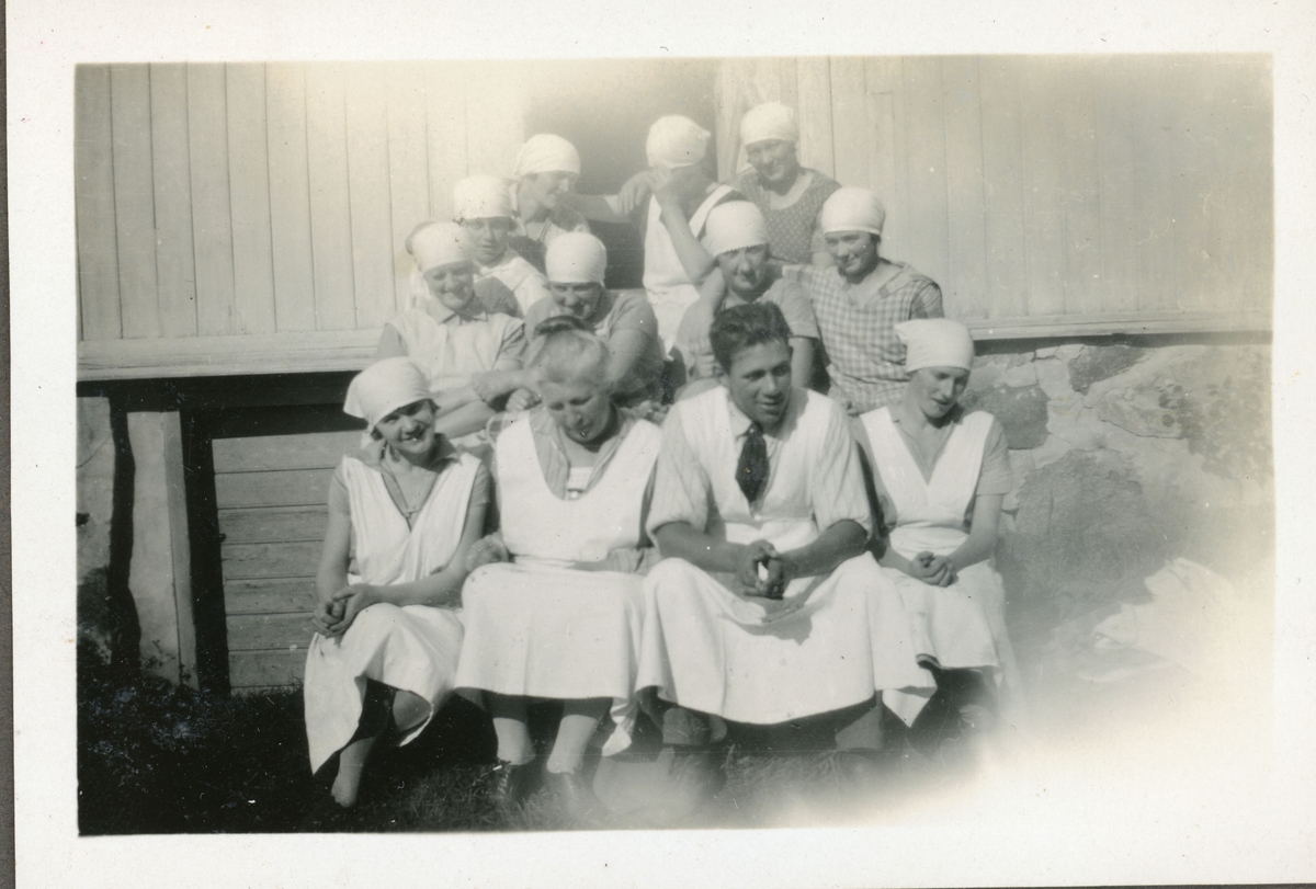 Elever fra husmorskolen på Skansgården, Kongsvinger, 1928. Ti unge kvinner i forklær og tørklær og eldre kvinne og yngre mann, også i forklær, sittende i uthustrapp.
