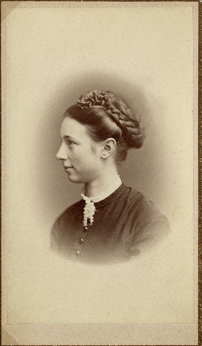 Porträttfoto av en kvinna i mörk klänning med liten vit ståkrage. Vid kragen syns en "brosch" (?). 
Bröstbild, profil. Ateljéfoto.