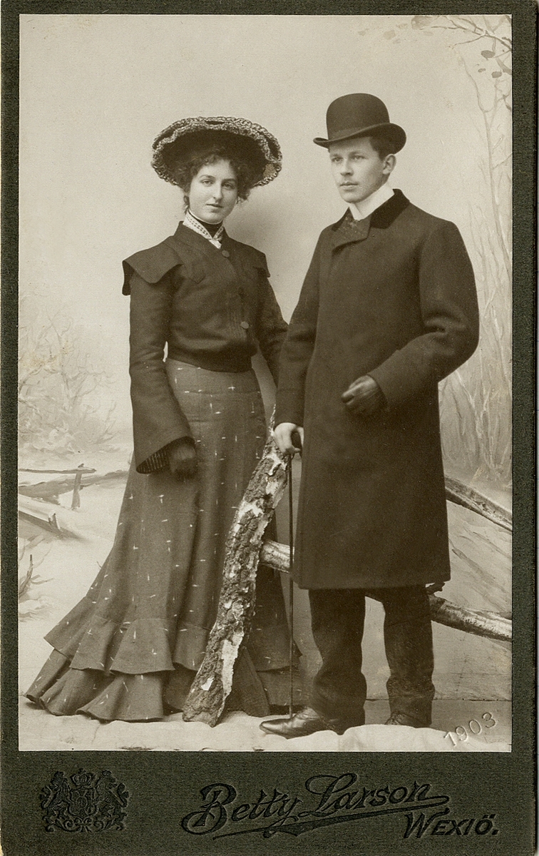Porträttfoto av ett ungt par. Hon är klädd i mörk mönstrad kjol med volanger, jacka, hatt och handskar. Han är klädd i mörk kavajkostym, paletå med sammetskrage, hatt och handskar. I ena handen skymtar en promenadkäpp. I nedre högra hörnbet syns inpräglat årtal: "1903".
Helfigur. Ateljéfoto.