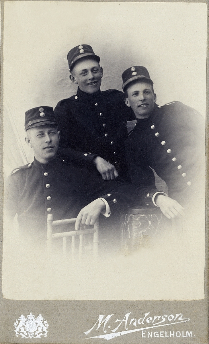 Porträttfoto av tre glada unga män i militäruniform. 
Midjebild, halvprofil. Ateljéfoto.