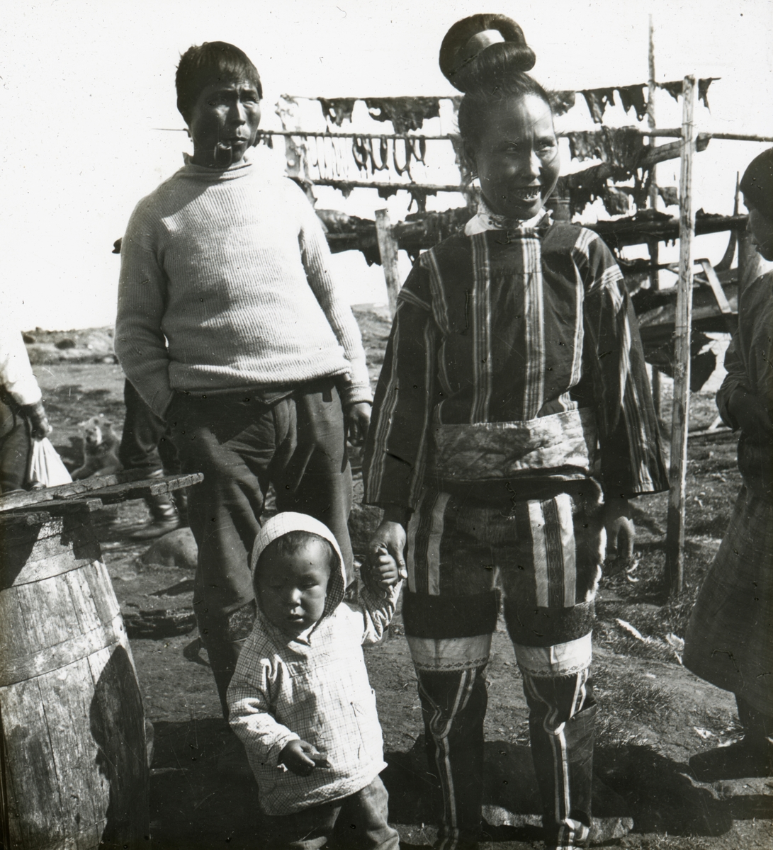 Motiv av en grønlandsk familie foran et stativ med tørket fisk.