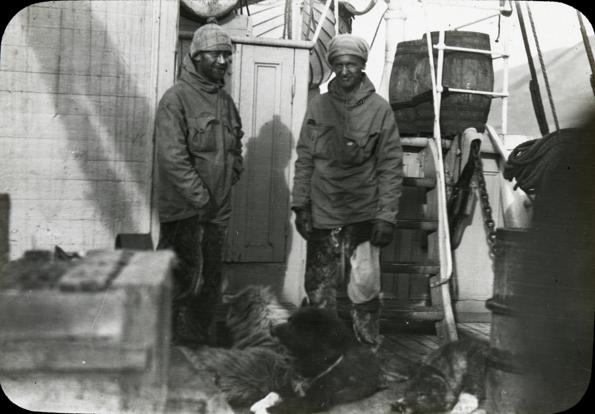 To menn ombord i ei ishavsskute. På dekk foran dem ligger noen grønlandshunder og hviler.