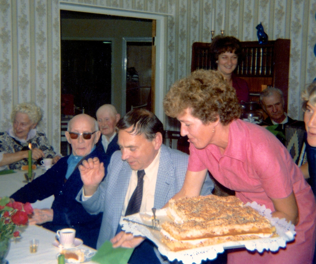 Kokerskan Ingela Sjöberg serverar tårta under en anhörigträff på Brattåshemmet 1979. Från vänster ses Knut Hagberg (i mörka glasögon), Alfred Mattsson (skymtar i bakgrundens mitt) samt Thure Bjerrhede (Knuts släkting från Vommedal). Relaterat motiv: A1892.