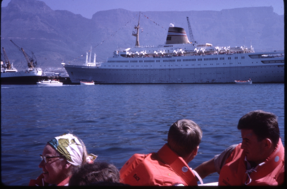Mennesker iført flytevester, med 'M/S Sagafjord' og flere livbåter i bakgrunnen. 'Sagafjord' Around The World via Africa Cruise 1966.