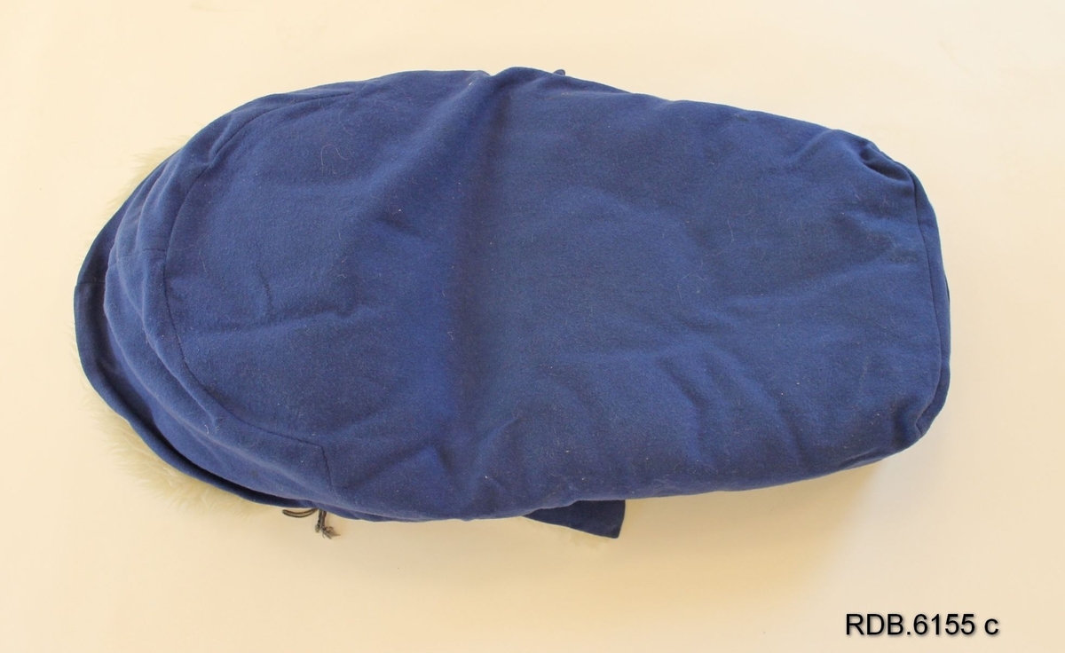 En babypose i blått ullstoff med kvitt saueskinnsfôr. Klaff foran som kan brettes ned og lukkes med 4 kvite knapper og hemper. Påsydd hempe på fotenden. Posen se hjemmesydd ut.