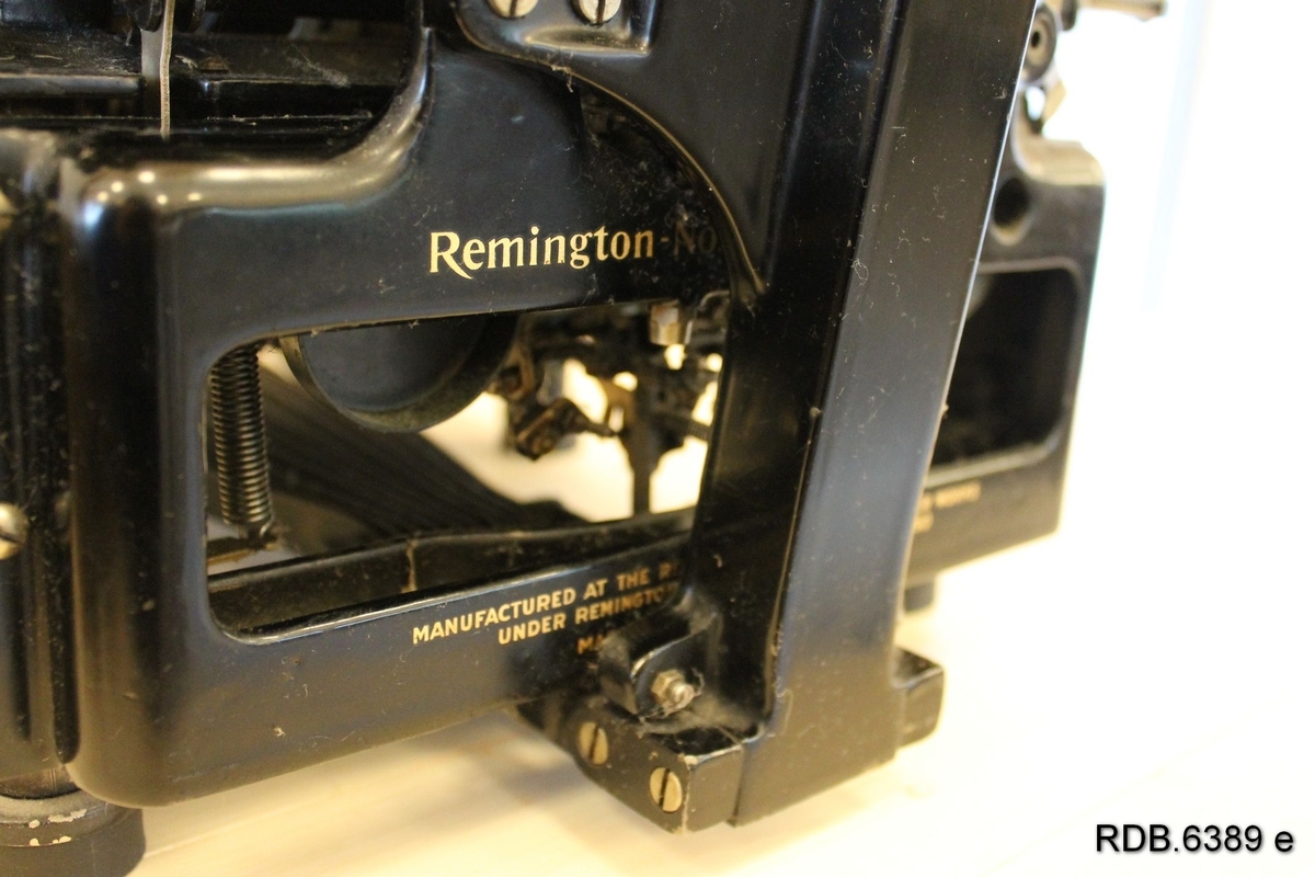 Svart skrivemaskin av merke Remington. Tastene er kvite med svarte bokstaver. På siden av skrivemaskinen har giveren limt på et bilde av og opplysninger om eier og bruker av maskinen.