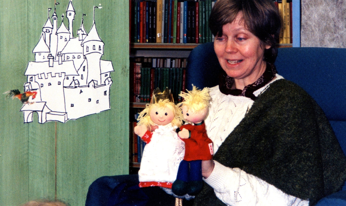 Sagostund för barn med Ingrid Hansson på Kållereds bibliotek 1990-tal. Till vänster ses en illustration av ett slott. Ingrid håller två dockor i händerna, varav det ena är en prinsessa.