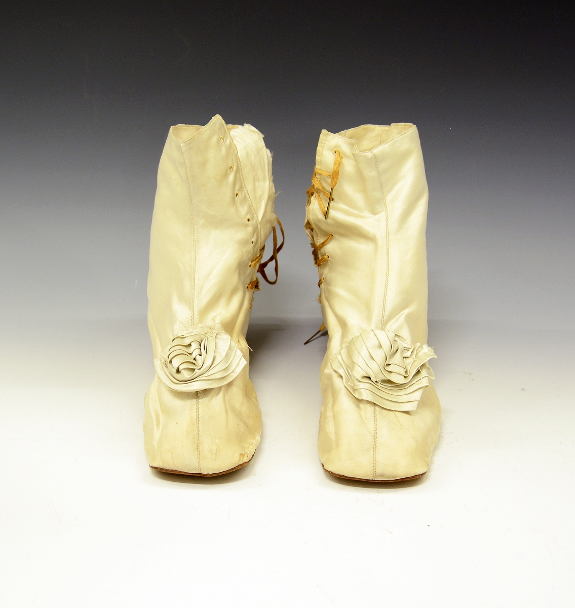 Silkestøvler. Hvite silkestøvler med høyt skaft og snøring. Sløyfe på tå og liten hæl. Silkestøvlene tilhører empirkjole TGM-BM.1923-24:068.