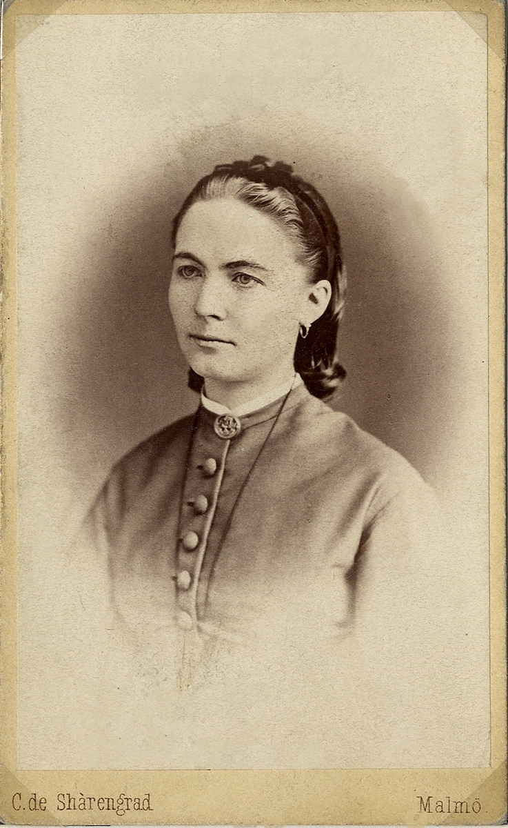 Porträttfoto av en kvinna i klänning med knappar och liten vit ståkrage. Vid kragen syns en brosch. 
Bröstbild, halvprofil. Ateljéfoto.