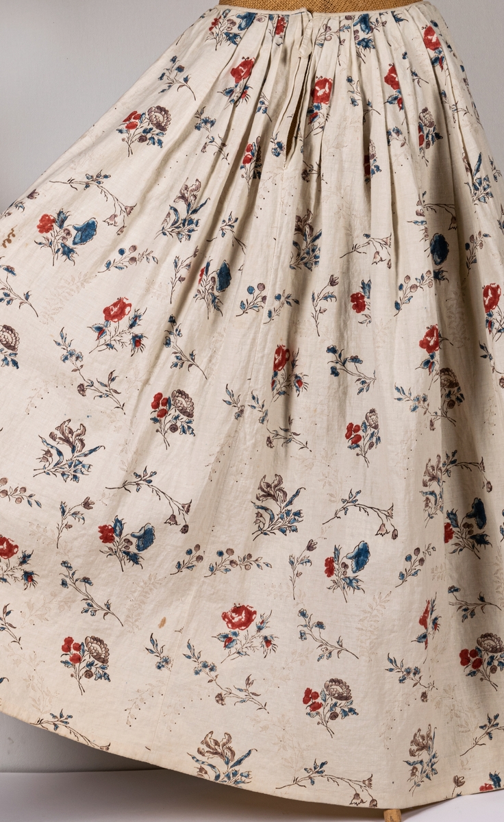 Tvådelad klänning, kjol och jacka, i naturfärgat linne med handtyckt blommönster i rött, brunt och blått. Klänningen är från 1700-talet.
a) jacka, urringad, knäppning fram, långa ärmar. Fodrad med naturfärgat linne. Lite svängd nedtill.
b) kjol, hellång, ofodrad, lagda vecka och smal linning.
