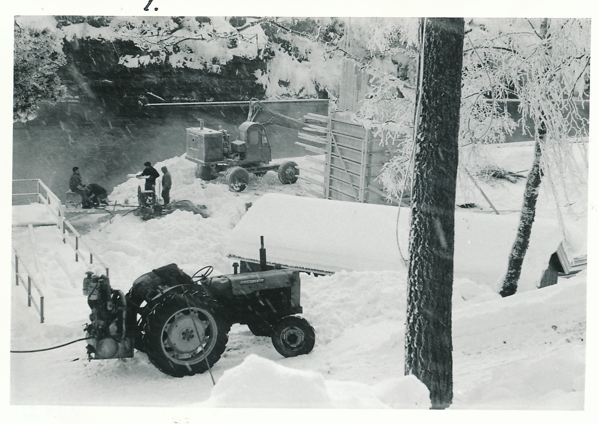 Anleggsarbeid i snøen ved båthuset nær Randselva på Kistefoss. Tre arbeidere, traktor og gravemaskin synlig, i tillegg til en konstruksjon under oppførelse der gravemaskinen står.
