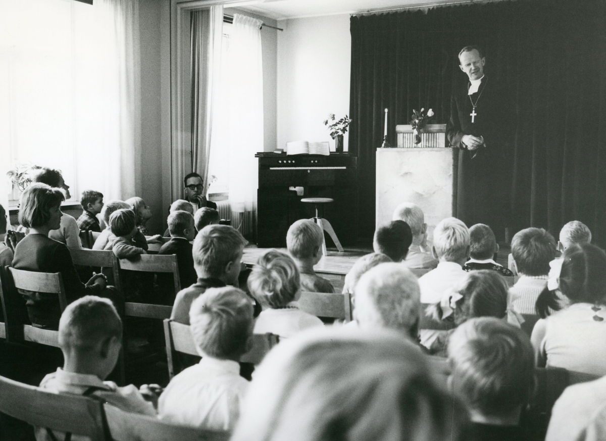 På podiet står biskop Bo Giertz som är på besök i Stretereds Lilla skola (Kållereds Folkskola) i samband med Biskopsvisitationen år 1966. Framför honom sitter elever.
