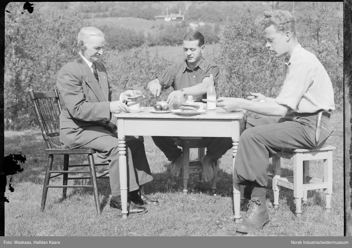 Tre menn spiser mat rundt bord utendørs. Brødmat og melkeflaske på bordet.