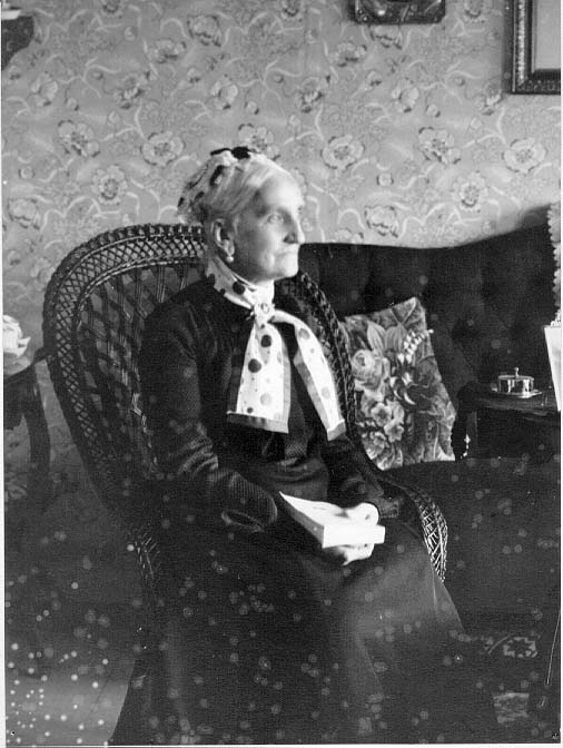 Fru Charlotte Fahnehjelm sittande i en korgstol med hög rygg i en hemmiljö. En scarf med kulörta prickar i olika storlekar runt halsen. Hon håller en bok i knät.
