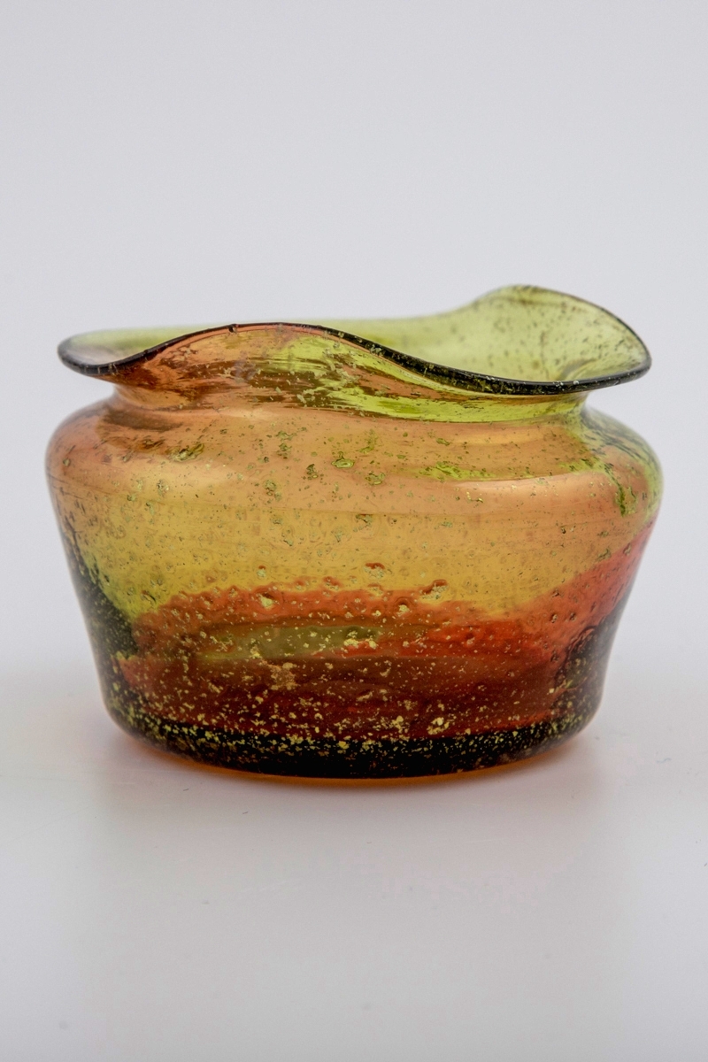 Liten vase i brungrønt, gjennomskinnelig glass med rødlige partier. Lav og rundt korpus med bølgende munningskant. Glassmassen har innlagt bladsølv. Puntemerke på vasens underside.