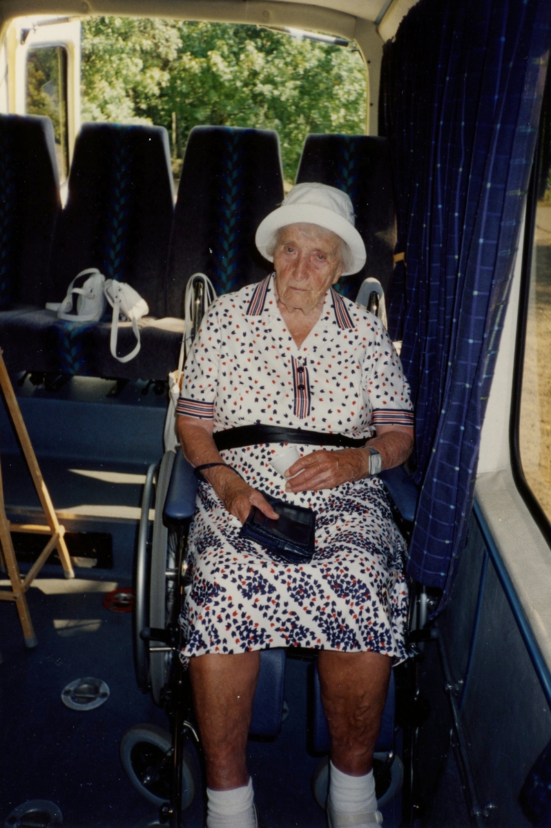 Boende från Brattåsgårdens äldreboende är på bussutflykt till Hembygdsgården Långåker, 1990-tal. Hulda Olsson, iklädd vit hatt och svart/röd-mönstrad vit klänning (1993 - 2000). Hon sitter i en rullstol som står i bussen. Hulda var född i Vommedal Östergård "Skräddarns".