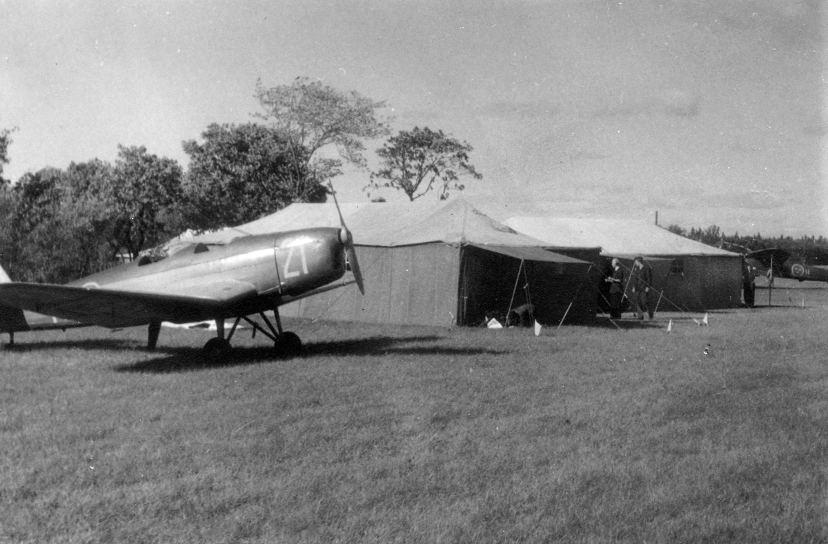 Flygplan Sk 15 uppställd på ett flygfält bredvid tälthangarer, 1940-tal. I bakgrunden syns ett par flygmekaniker och flygplan Saab B 18.