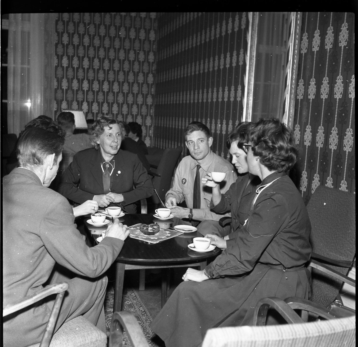 Män och kvinnor, några i scoutdräkt, sitter i grupper runt bord med kaffekoppar.