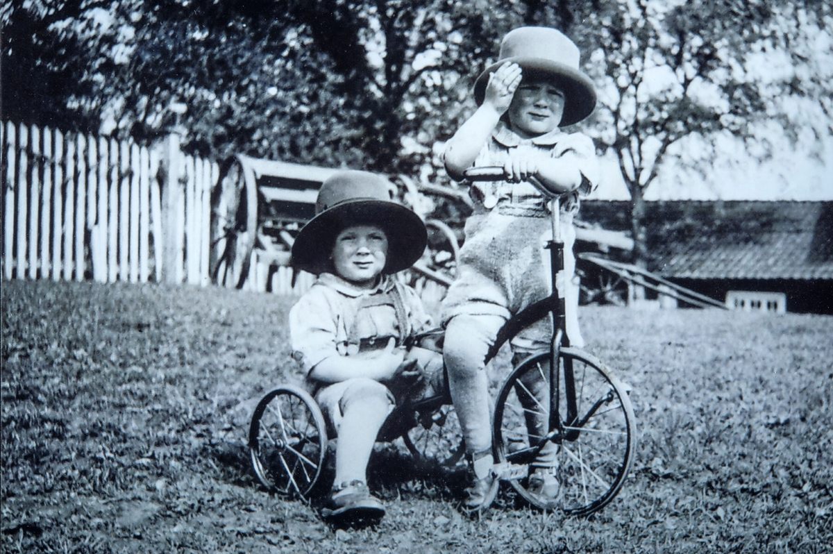 2 barn med trehjulsykkel. Tvillingene Thorstein og Terje Rustad, på Dælin Østre i Ottastad leker seg med trehjulsykkelen sin på gårdstunet på Dælin. Thorstein sitter på sete og Terje på bagasjen. Tvillingene er født i 1937.