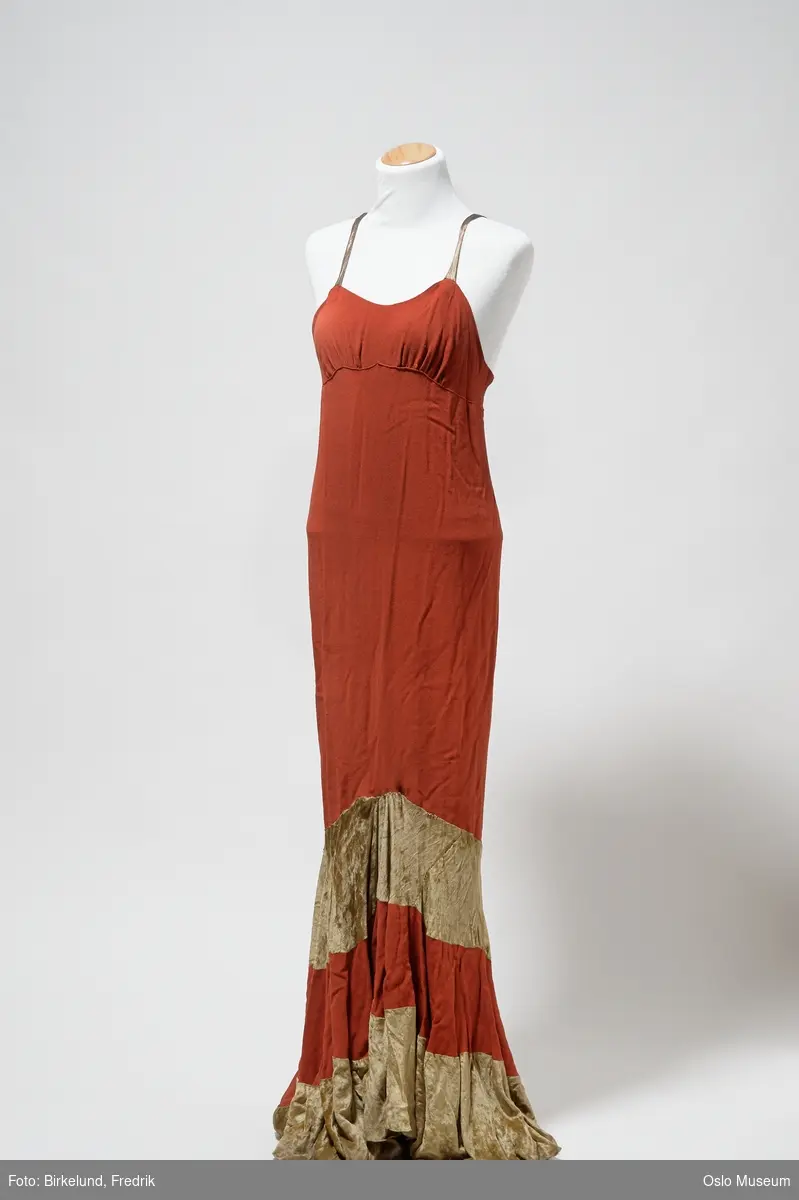 Lang rød kjole med skulderstropper. Nedre del av kjolens bakside er lengre enn kjolens forside. Nedre del av skjørt består av annehver rød og gullbeige tekstil. Lukkes med glidelås i ryggen.