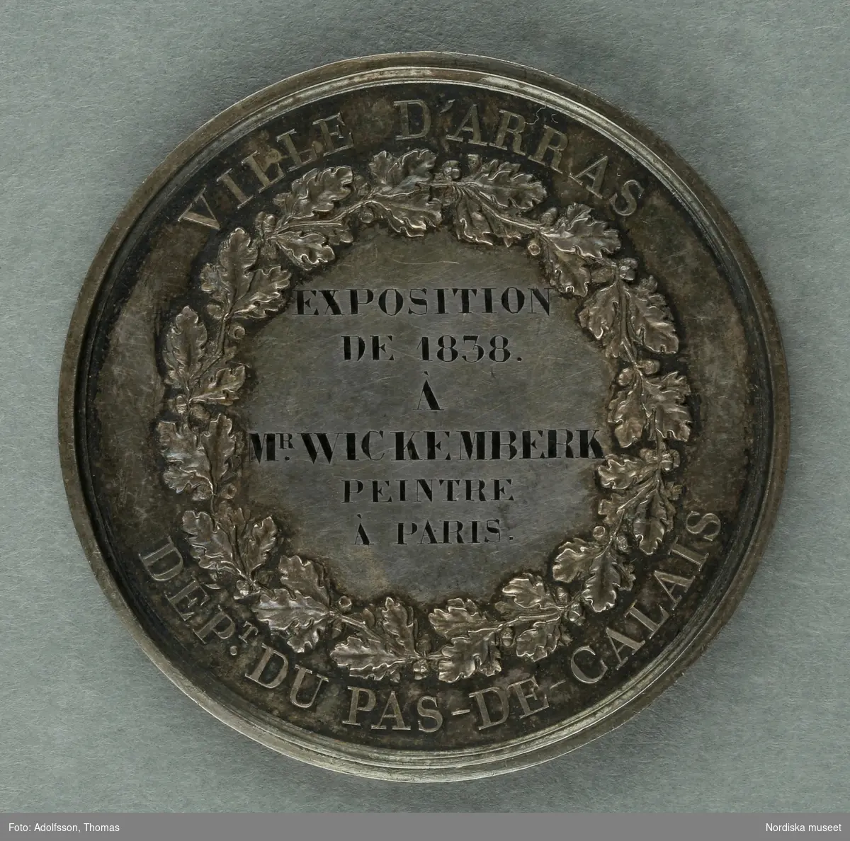Huvudliggaren.
"Medalj. I hvit metall. Åtsidan: 'Louis Philippe I Roi des francais'. Bröstbild v.o. Under bilden: 'BARRE F.T [t:et snett ovanför F.et]. Frånsidan öfverst: 'Ville d'Arras'. Inom en eklöfskrans: 'Exposition de 1838. A. M [efter m;et svårtytt tecken] Wickemberk peintre à Paris'. Nederst utom kransen: 'Dep .T [t:et ovanför punkten] du Pas-de-Calais.'. 
Diam lin.
G. fr basaren f. Skdin. etn. saml. [=för Skandinaviska etnografiska samlingens] byggnadsfond, gm. fru Olivecrona f. Roos, i apr. 1877."