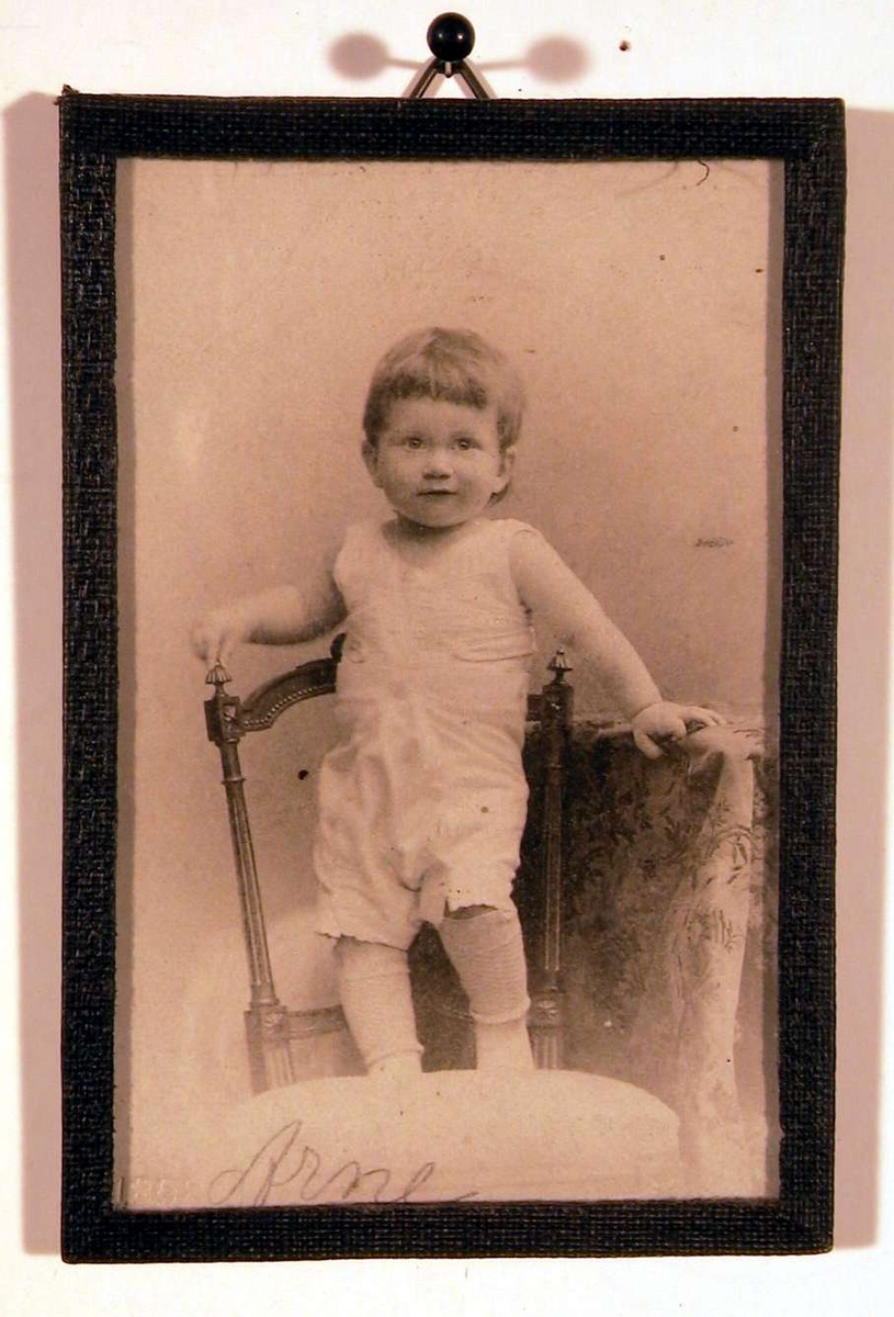 Barneportrett, liten gutt i hvitt undertøy stående på stol, venstre hånd støtter på bordkant. Han ser direkte på betrakteren.