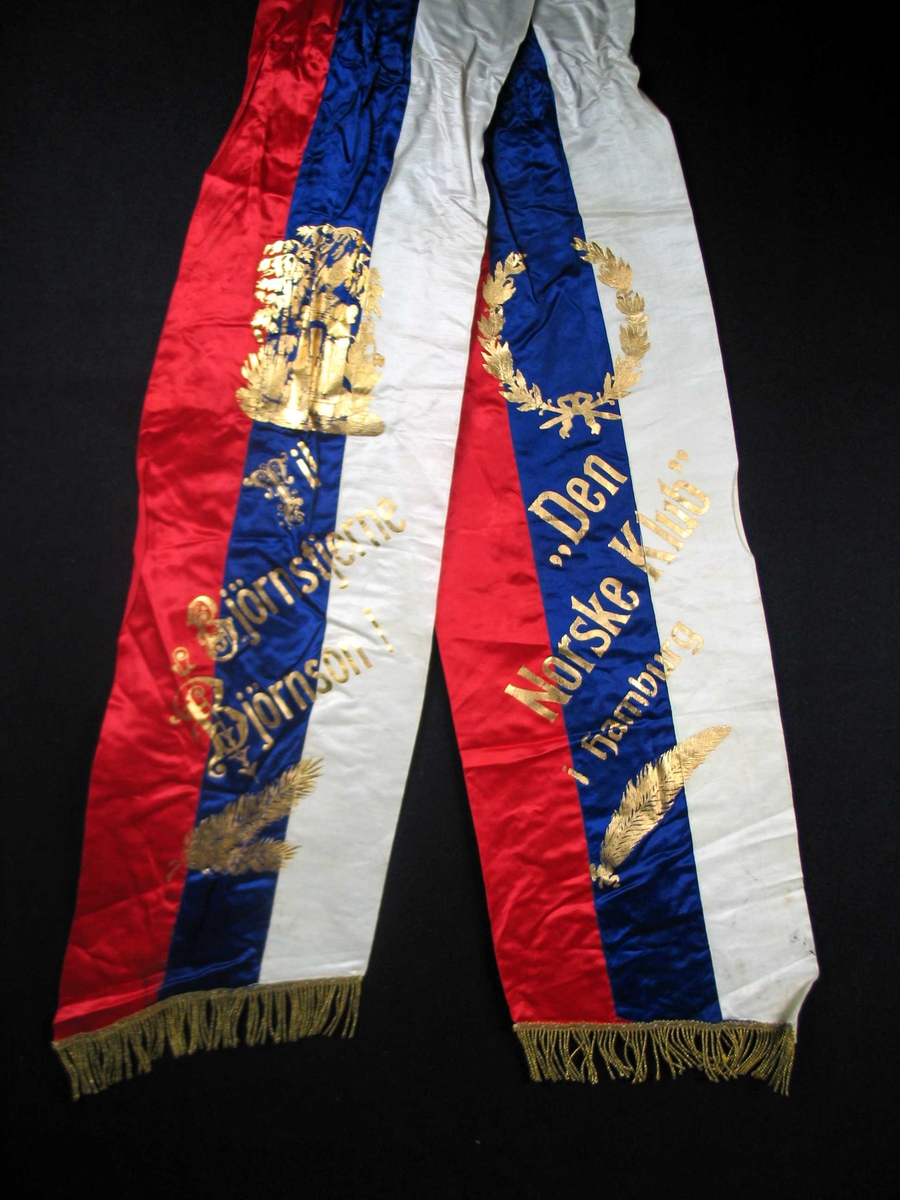 Begravelsessløyfe i rød, hvit og blå silke med frynser og tekst i gull.