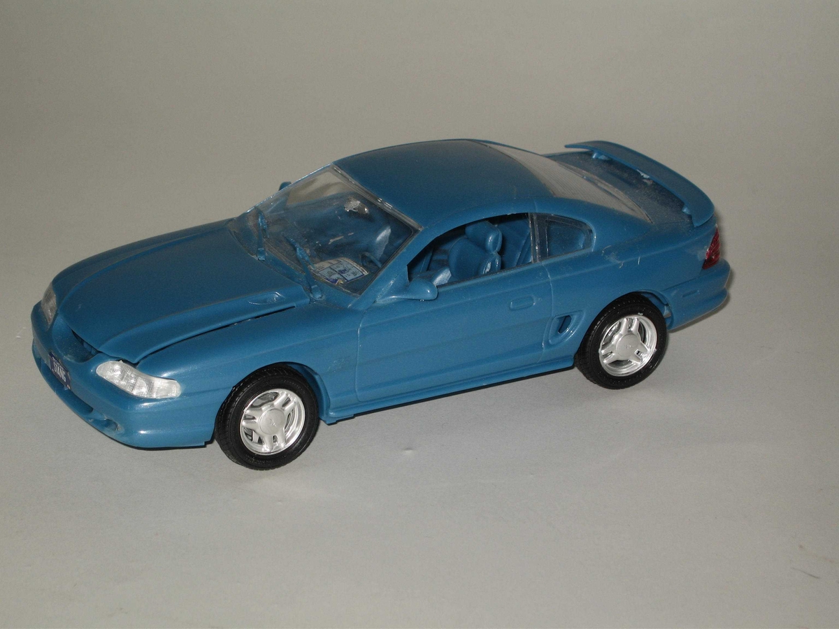 Leketøysbil som modellbil satt sammen av mange deler, hvorav karosseri og understell er laget av blå plast, vinduer og framlykter av klar plast, baklykter av rød plast, dekkene av svarte plast og felgene av kromfarget plast. Bilen er en modell av en Ford Mustang.
