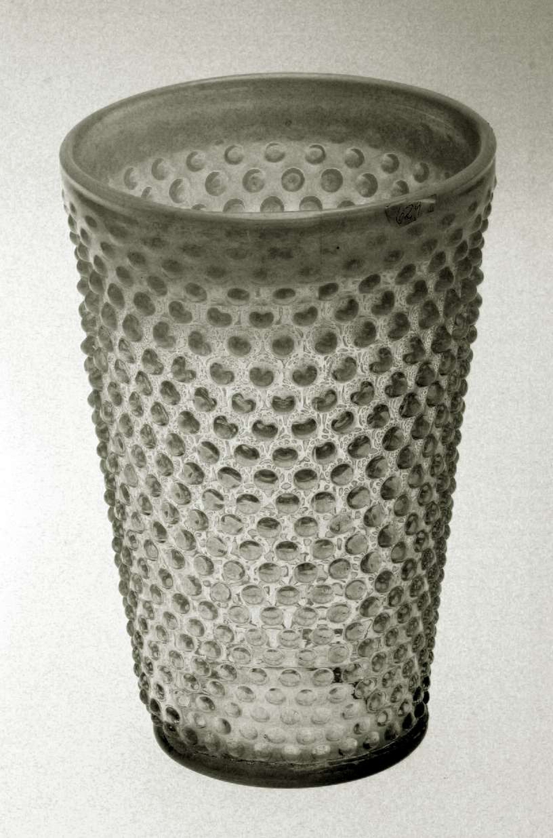Blomstervase med konisk form. Den er i glass og yttersiden er 'knoppete'. Kanten av vasen og 'knoppene' har en melkehvit farge. Vasen er sprukket.