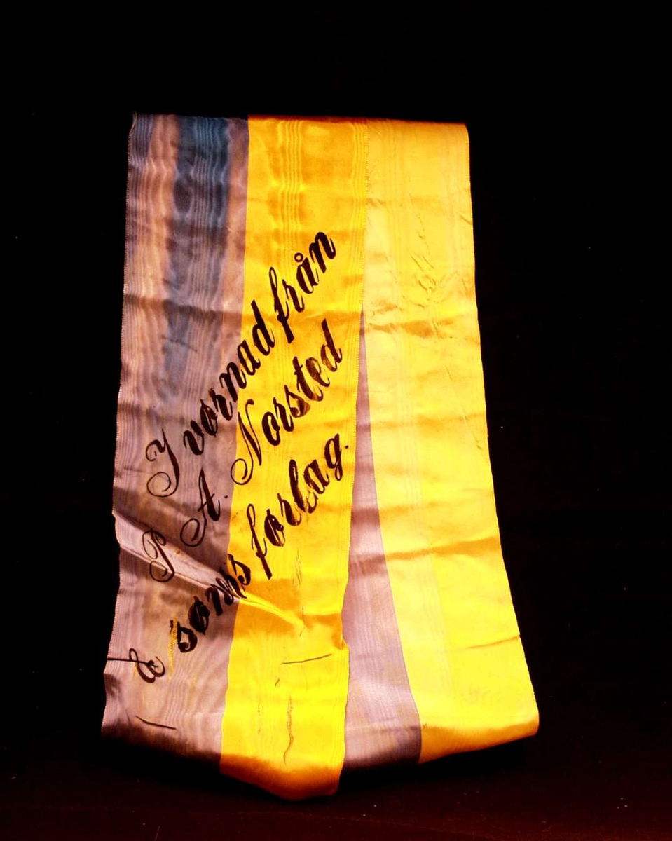 Bånd i gul og blå silke. Båndet er delt i to. På båndet står det: I vørnad från P. A. Norsted & søners førlag.