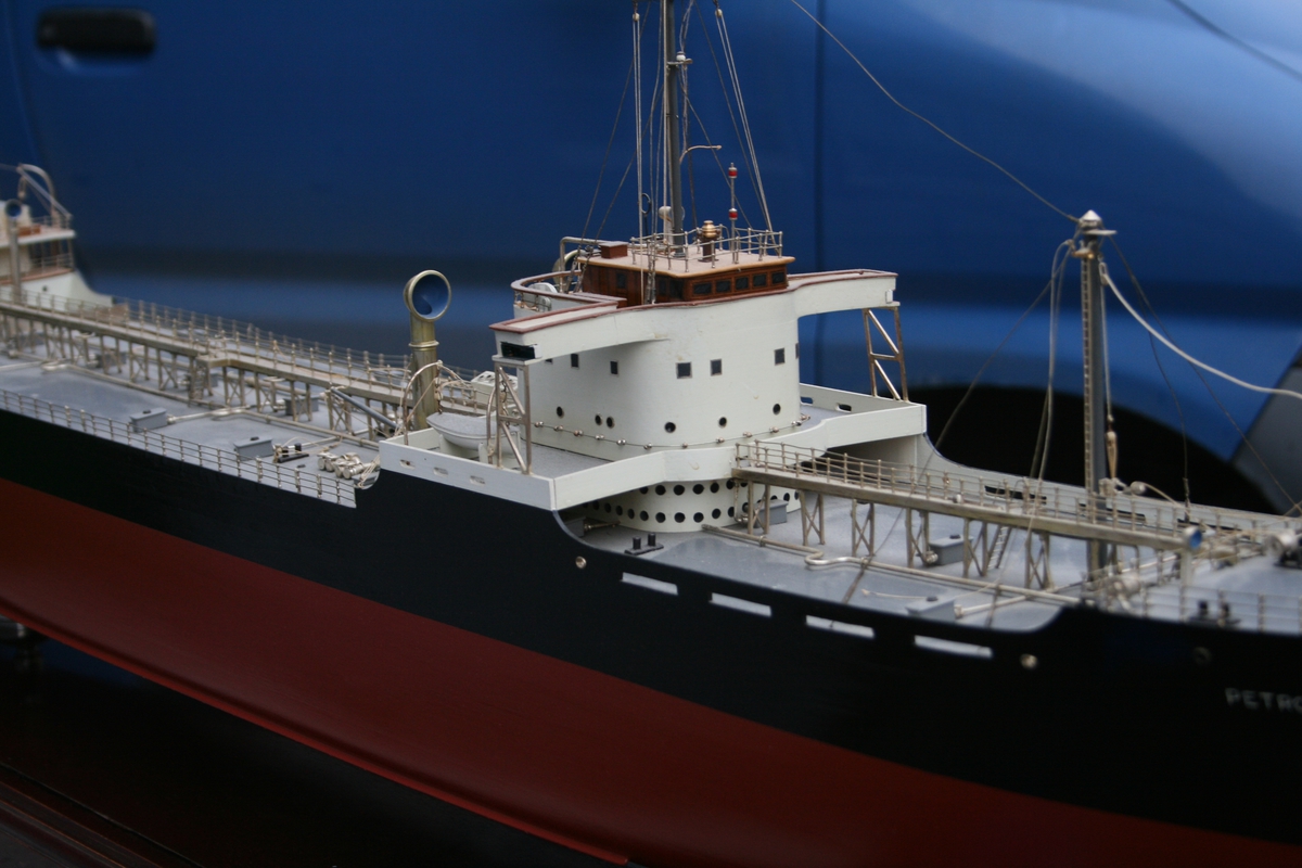 Skipsmodell, tankbåt, i monter. Pipemerke: Blått bånd rundt sort pipe, hvit rombeformet felt med rød bokstad H.  Monteren er beiset i palisanderfarge og lakkert.
