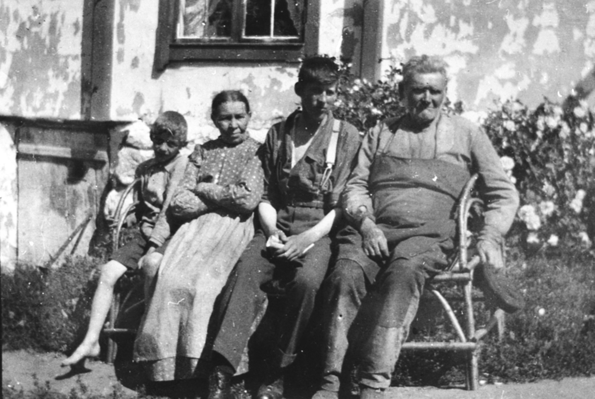 4 personer sitter utenfor huset i Sundby lille, Skredderbakken, Helgøya. Fra venstre Asbjørn Solmund Aas f. 1912, Marit Jensdatter (1855-1939), Leif Sæhlie, Johannes Akselsen (1844-1926).