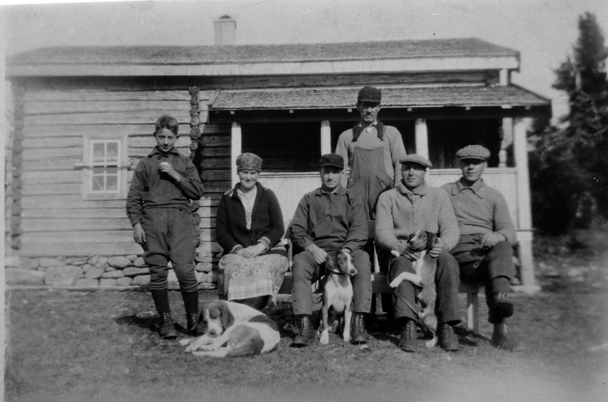 6 personer foran hytte, jegere. Sittende til høyre er Aksel Johansen, til venstre med hund er Ole Haugli.