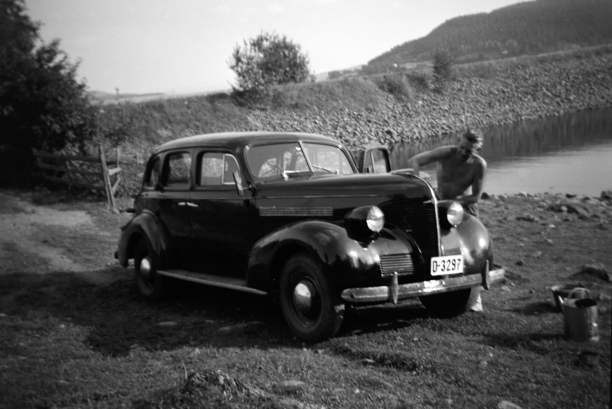 Bilvask. Arkitekt Rolf Prag vasker bil utenfor hytta si i Kvarbergvika i Brumunddal. Personbil D-3297. Chevrolet 1939. Ca. 1950. Fyllinga til den gamle Framnesbrua i bakgrunnen. 
