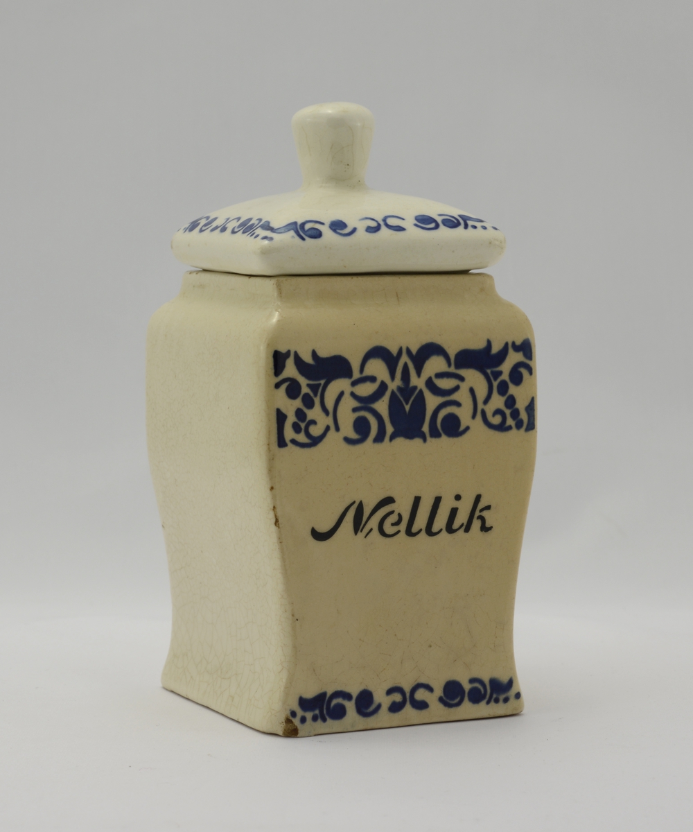 Hvit kvadratisk krydderkrukke i hvit keramikk, med påskrift "Nellik" i svart. Blank glasur. Organisk ornament i blått på krukkens front; et bredt belte over "Nellik", et smalt belte nederst på krukkens front. Lokket har den samme type ornament langs randen, og har en knott. Krukken er ganske gulnet, hvilket gjør at fargeforskjellen mellom lokk og krukke er ganske stor.