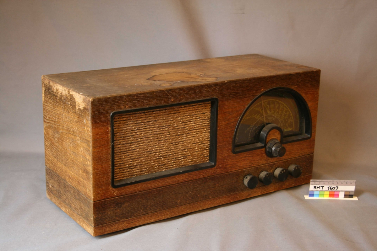 Rektangulær brun radio. Forside: skala med fem funksjons- og søkeknapper. Høyttaler med trekk av tekstil. Bakside: hele bakplaten mangler. 