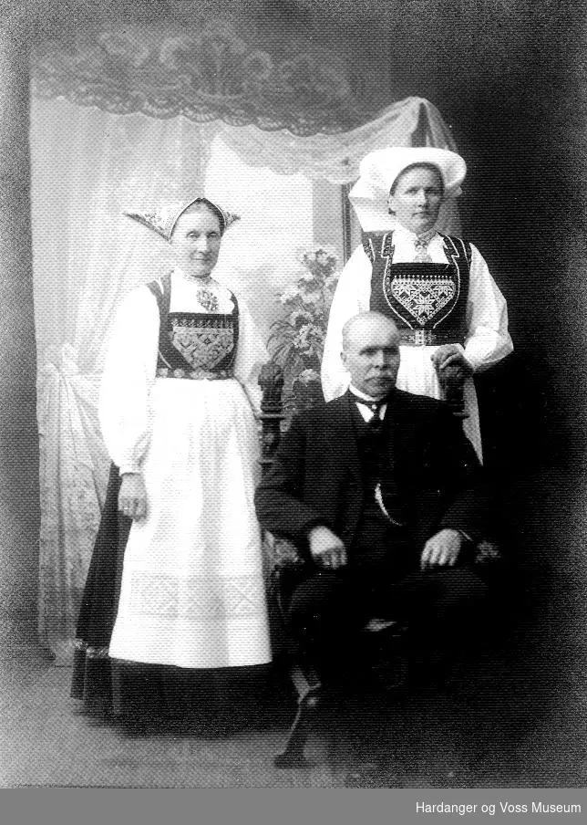 familiegruppe i fotoatelier, to kvinner i bunad, og ein mann i dress