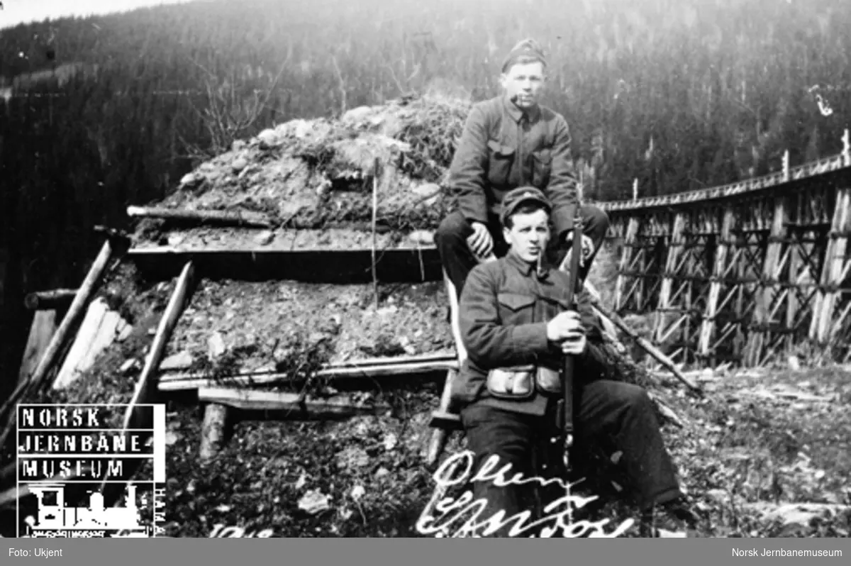 To militære foran ei torvhytte med Drøia bru i bakgrunnen